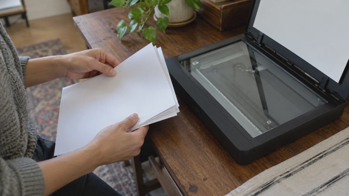Online Document Vault: Practical check Hands checking a stack of pages beside an open flatbed scanner.
